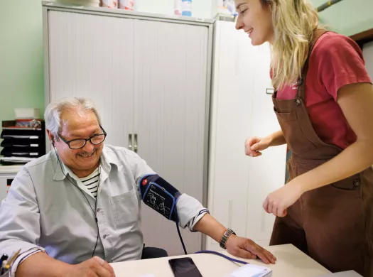 Een man zit aan een bureau terwijl een jongere vrouw zijn bloeddruk meet